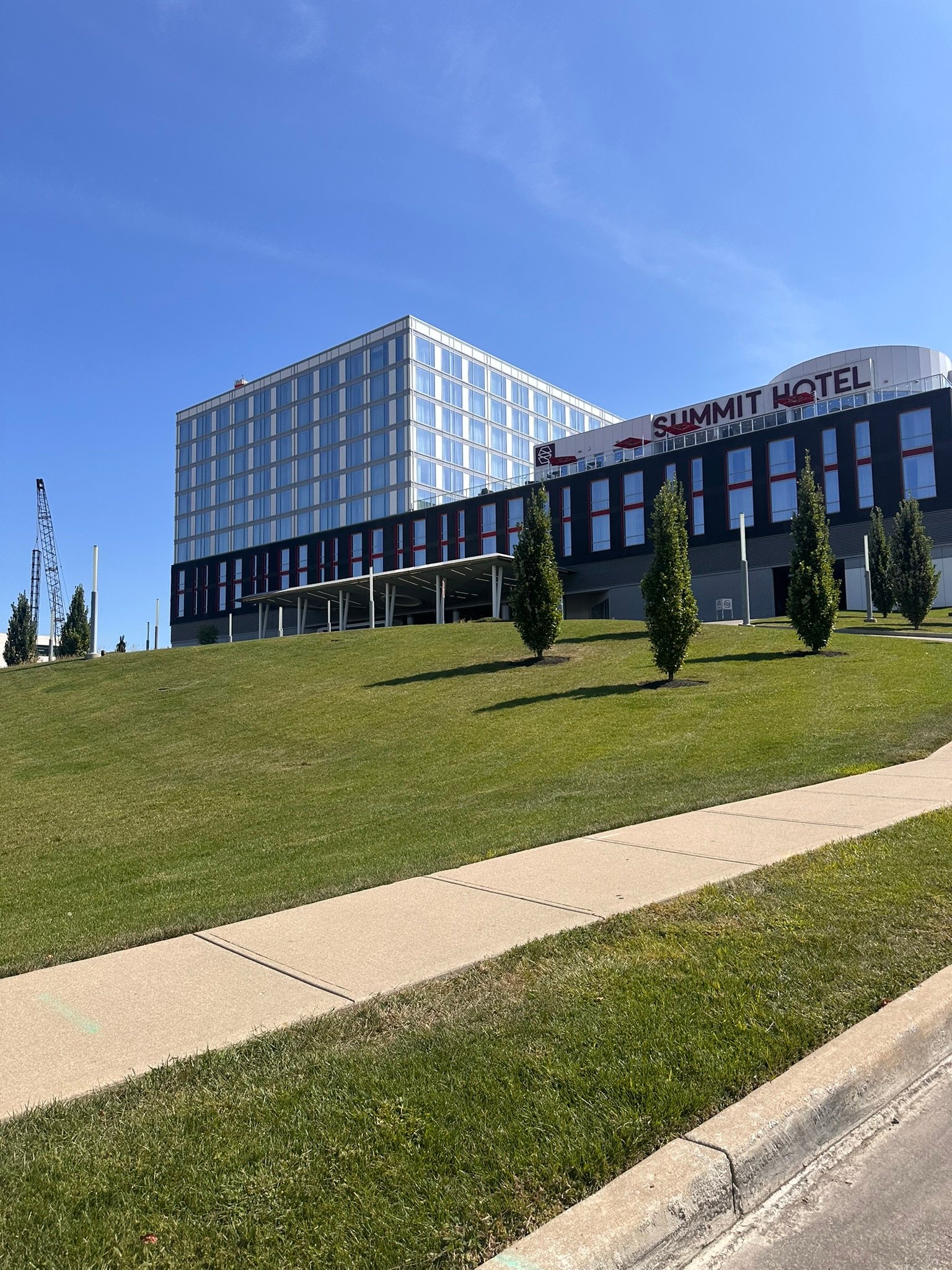 A modern hotel building labeled Summit Hotel sits atop a grassy hill under a clear blue sky, with a sidewalk and young trees in the foreground.