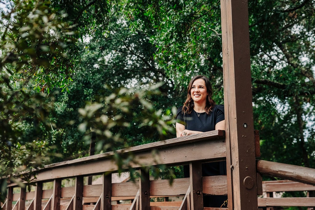 A woman in a black dress stands and smiles on a wooden bridge surrounded by green trees.